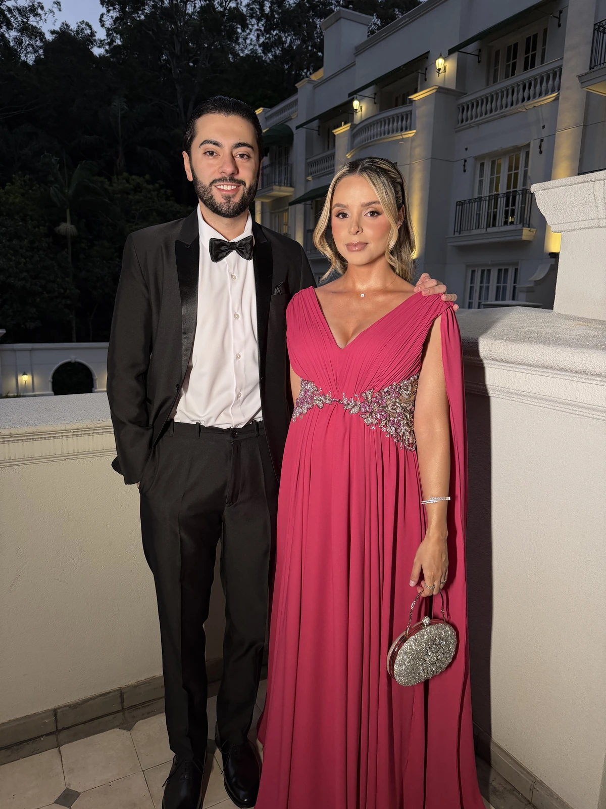 Casal em traje de festa ao ar livre, com vestido rosa e smoking preto.